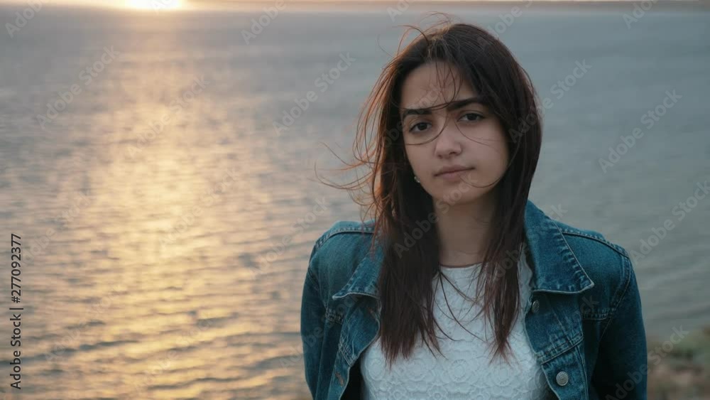 Portrait of young beautiful brunette lady standing on the mountain near the sea with strong wing waving her hair at sunset in slow motion Portrait of young beautiful brunette lady standing on the mountain near the sea with strong wing waving her hair at sunset in slow motion