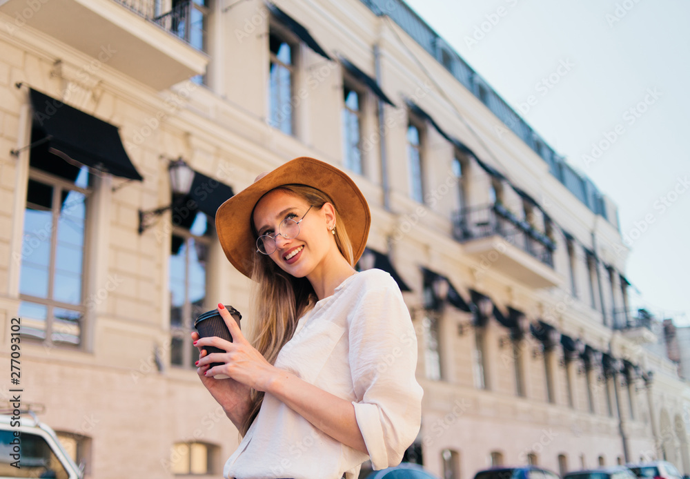 Young casual style woman in felt hat holding cup of coffee on the go against the background of urban architecture