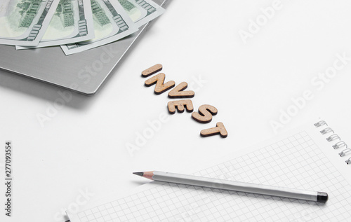 Creative business concept. Laptop, notebook, pencil, dollar bills on white background with the word invest of wooden letters, minimalism