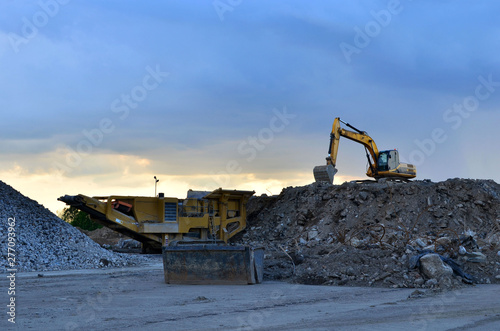 Heavy excavator working in quarry on a background of sunset and blue sky. Mobile jaw stone crusher by the construction site. Crushing old concrete wastes and subsequent cement production