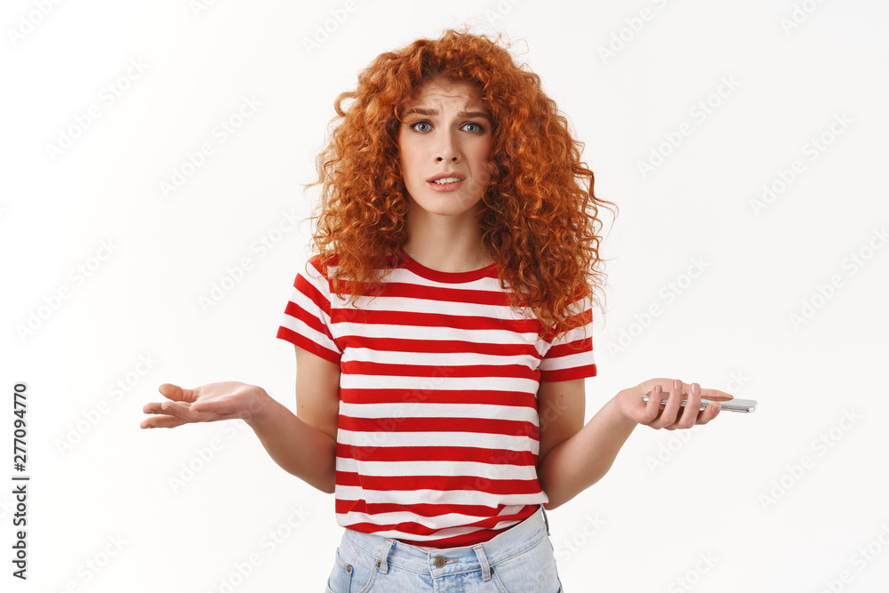 Obraz premium Confused uncertain stylish beautiful redhead curly girlfriend shrugging dismay puzzled holding smartphone hands spread sideways stare questioned trying understand who spam mailbox
