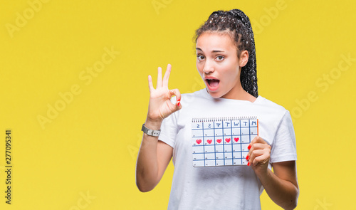 Young braided hair african american girl holding menstruation calendar over isolated background doing ok sign with fingers, excellent symbol