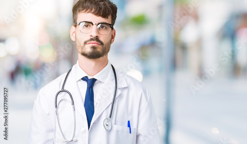 Young doctor man wearing hospital coat over isolated background Relaxed with serious expression on face. Simple and natural looking at the camera.