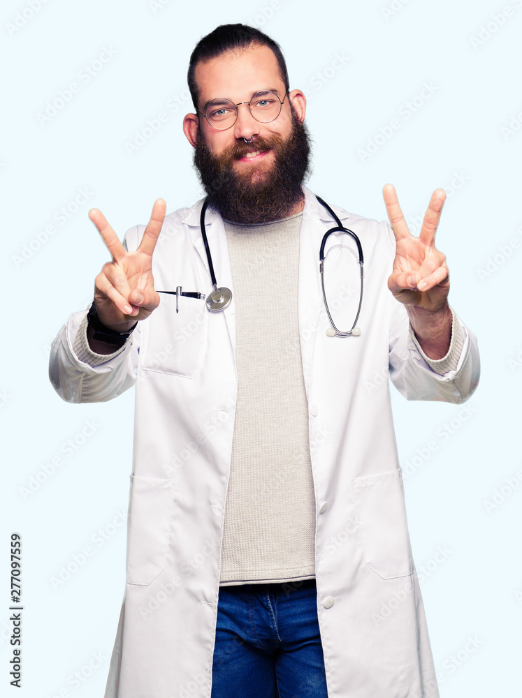 Young blond doctor man with beard wearing medical coat smiling looking to the camera showing fingers doing victory sign. Number two. Young blond doctor man with beard wearing medical coat smiling looking to the camera showing fingers doing victory sign. Number two.