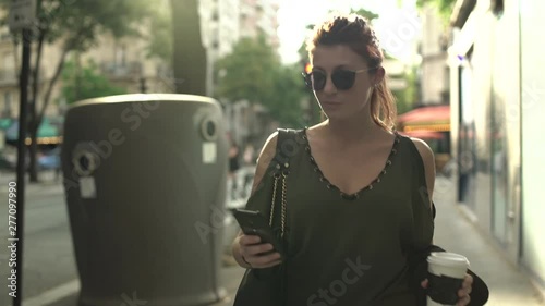 Attractive smiling caucasian woman with sunglasses, freckles, piercings and red hair writing text message to boyfriend on her smartphone and drinking coffee walking through the street in Paris. 4K.
