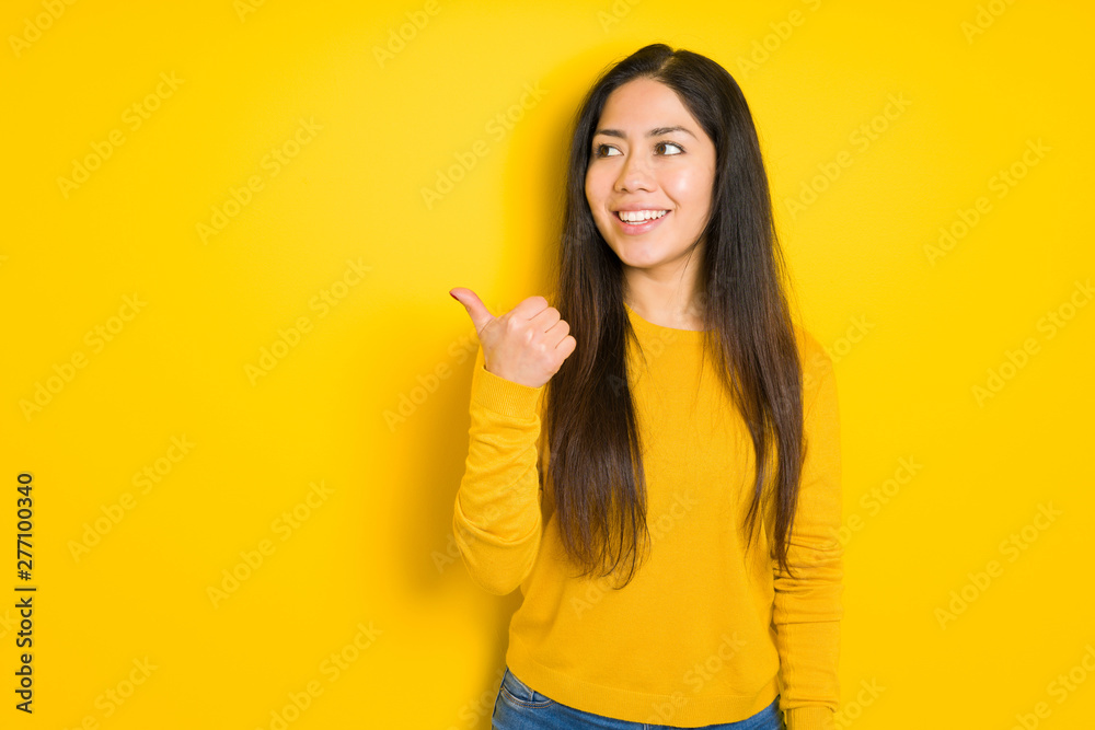 Beautiful brunette woman over yellow isolated background smiling with happy face looking and pointing to the side with thumb up. - obrazy, fototapety, plakaty 