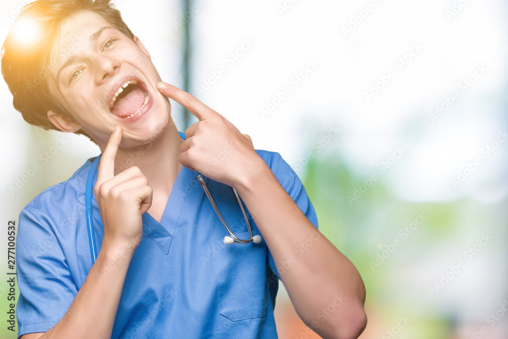 Young doctor wearing medical uniform over isolated background smiling confident showing and pointing with fingers teeth and mouth. Health concept. Young doctor wearing medical uniform over isolated background smiling confident showing and pointing with fingers teeth and mouth. Health concept.