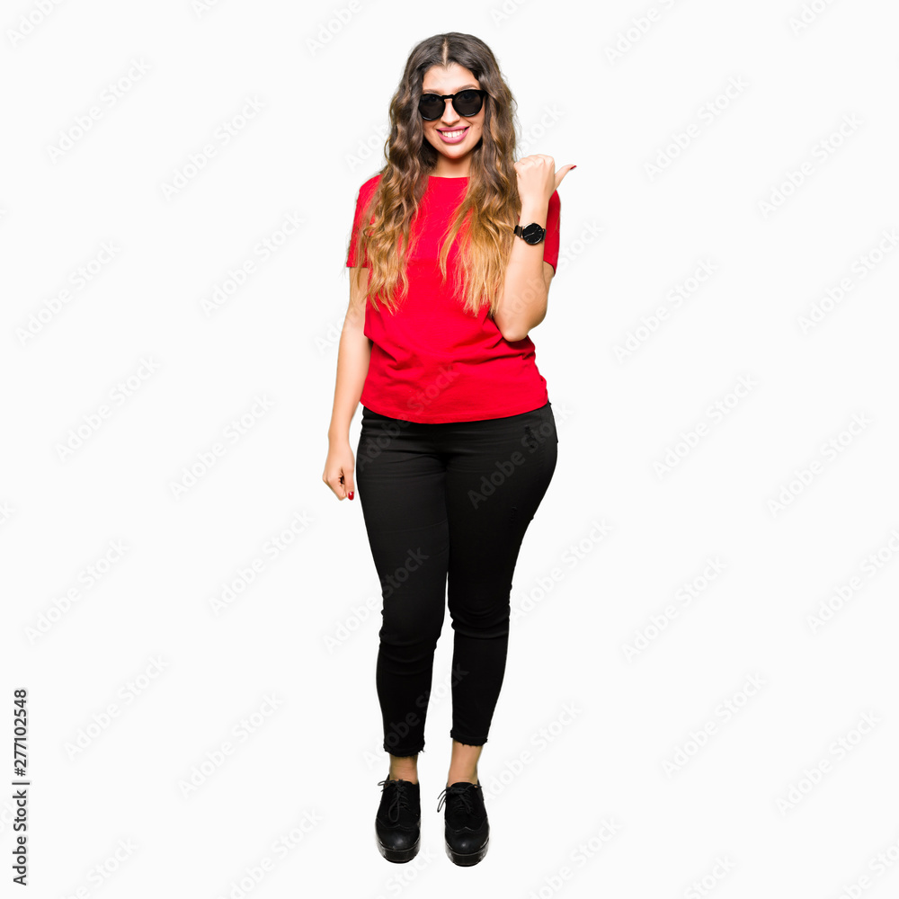Fototapeta premium Young beautiful woman wearing red t-shirt and sunglasses smiling with happy face looking and pointing to the side with thumb up.