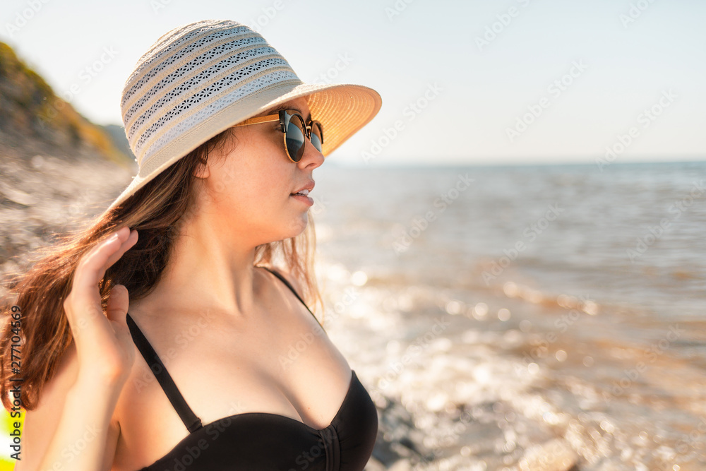 A young attractive woman in a hat and sunglasses looks into the distance. The sea in the background. Tint