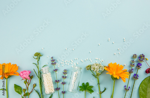 Flat lay view homeopathic medicine pills in jars and spilled around on light blue background, decorated with fresh various herbs and plants, flowers. Homeopathy border background, lot of copy space.