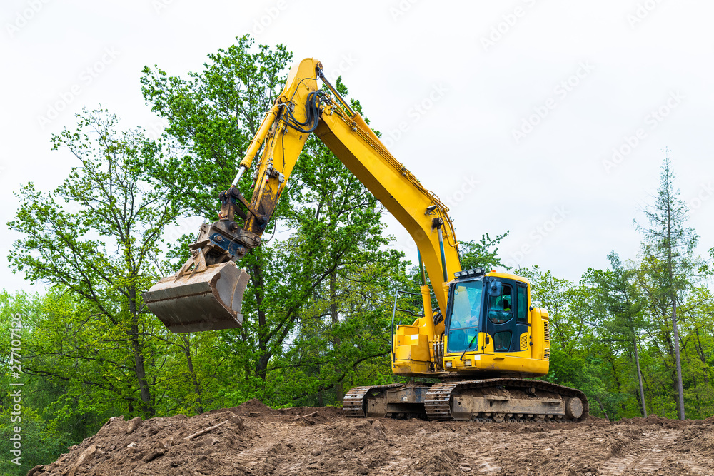Yellow belt excavator on construction site. Hydraulic earth mover. Backhoe loader. Rotating operator cabin, shovel and continuous tracks. Pile of soil, spring sky, green tree tops. Building industry. Yellow belt excavator on construction site. Hydraulic earth mover. Backhoe loader. Rotating operator cabin, shovel and continuous tracks. Pile of soil, spring sky, green tree tops. Building industry.
