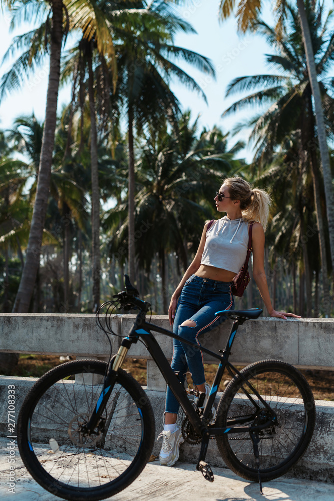 Naklejka premium Pretty woman with Bicycle on a background of palm trees forest. Young girl riding bike in Vietnam or Thailand tropical jungle with coconut palms. Summer holiday tourist activity. Active sports tourism