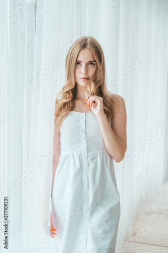 Girl in a light white dress posing by the window.