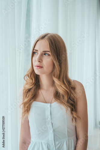 Girl in a light white dress posing by the window.