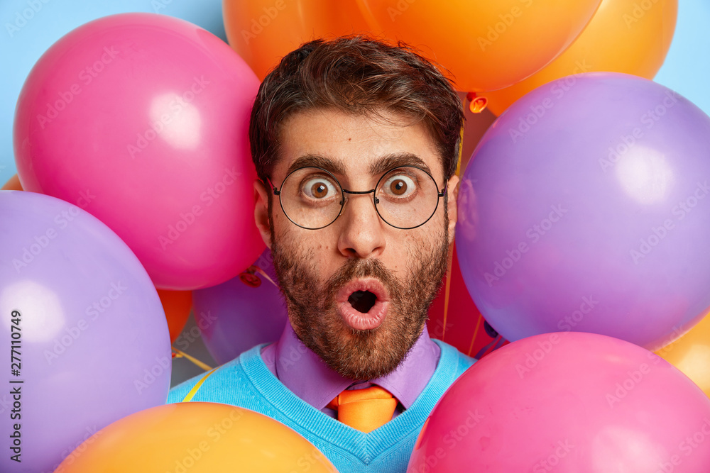 Shocked party man wears optical spectacles, cannot believe eyes, being in stupor, reacts on holiday organisation, surrounded with colorful balloons. Festive event, emotions, human facial expressions - obrazy, fototapety, plakaty 