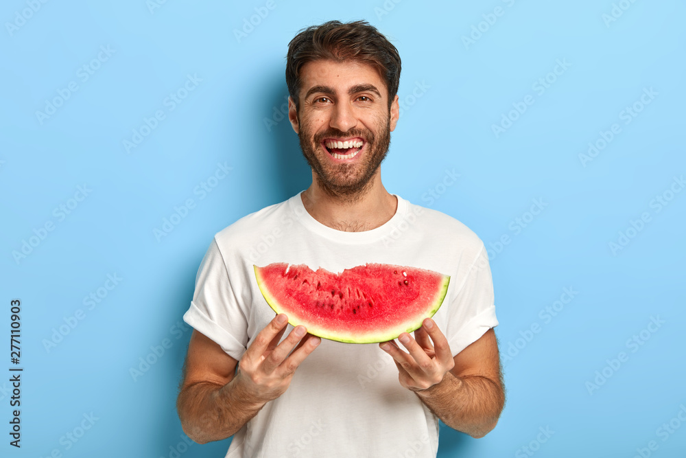 Waist up shot of smiling man enjoys summer day, holds slice of fresh ripe watermelon, wants to eat delicious fruit, wears casual white t shirt, isolated on blue background, has picnic during weekend - obrazy, fototapety, plakaty 