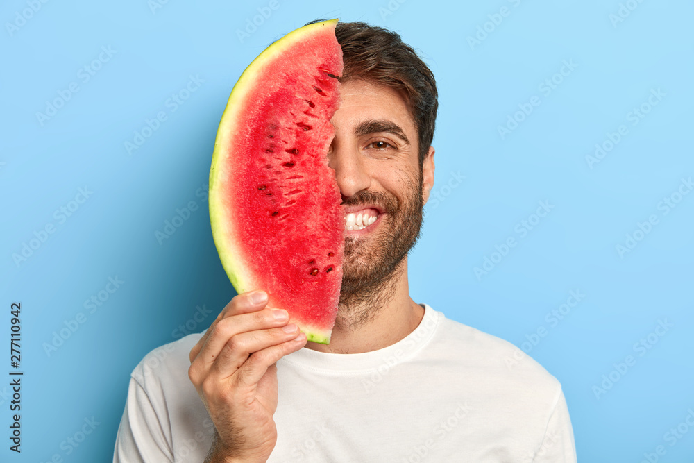 Taste of summer concept. Cheerful young man covers half of face with big slice of watermelon, has oraganic healthy nutrition, dressed casually, poses on blue background, enriches with vitamins. - obrazy, fototapety, plakaty 