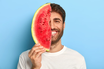 Taste of summer concept. Cheerful young man covers half of face with big slice of watermelon, has oraganic healthy nutrition, dressed casually, poses on blue background, enriches with vitamins.