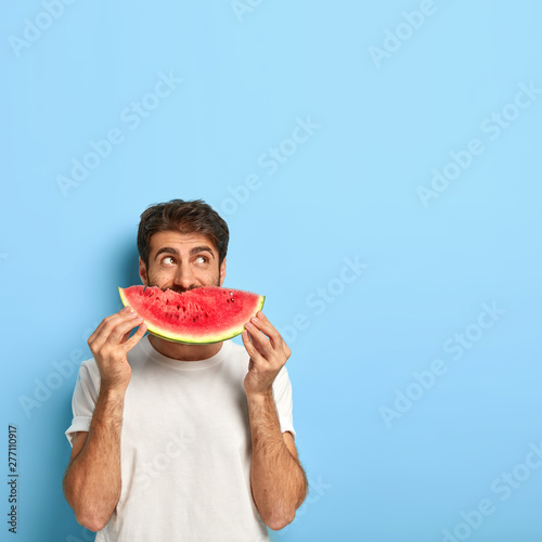 Vertical shot of attractive young man covers mouth with slice of red juicy watermelon  enjoys eating seasonal tropical fruit during summer period  wears white t shirt  poses indoor. What nice taste 