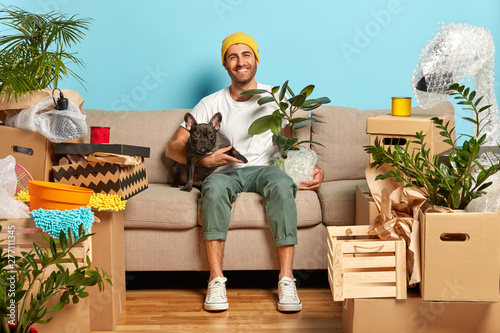 Indoor shot of delighted carefree house owner sits on couch and hugs pet, rests in own apartment, being in high spirit, celebrates move, has fun at home much household stuff around. Relocation concept