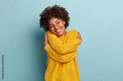Lovely happy African American woman daydreams, embraces herself, recalls romantic date, feels comfort, keeps hands across body sensually, has broad smile, wears yellow sweater, poses indoor. photo
