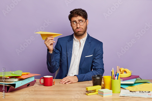 Portrait of young experienced entrepreneur thinks about main theses for business deal, being well dressed, holds paper plane, looks thoughtfully aside, drinks coffee, poses at desktop in cabinet photo