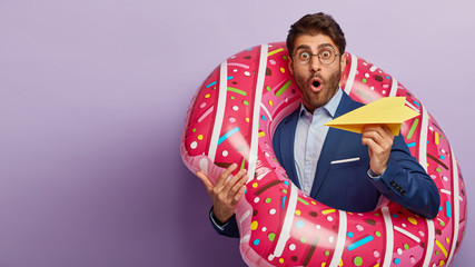 Back to work concept. Astonished director shocked summer holidays ended so quickly, holds paper plane, wears elegant clothing, stands inside of inflated swimring, isolated on purple blank wall