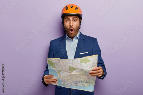 Emotional surprised engineer reads construction map, shocked to reloacte on new construction site, wears orange protective helemt, formal clothes, isolated on purple wall. Work and building concept photo