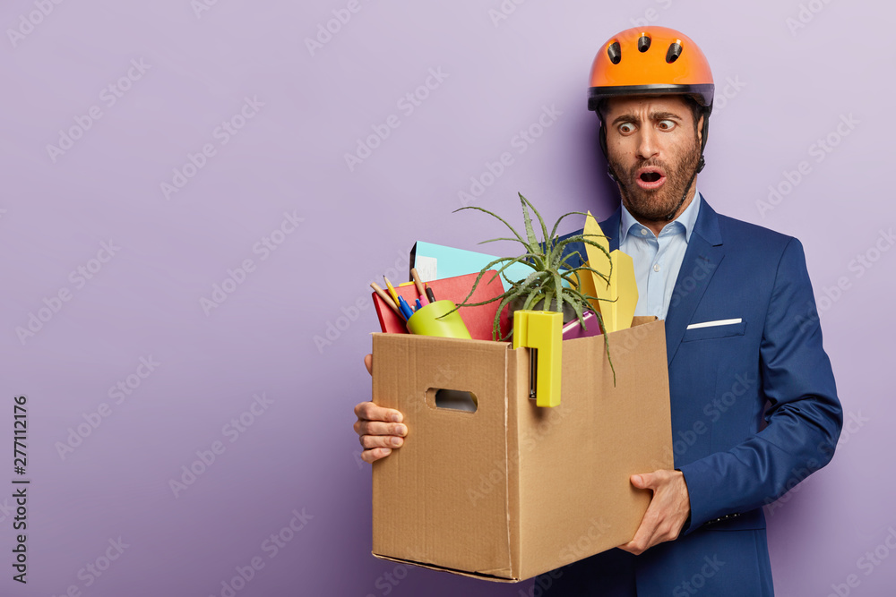 Shocked construction manager fired from work, holds cardboard box with office supplies, has surprised frustrated expression, dressed in elegant suit and protective helmet. Changing workplace Shocked construction manager fired from work, holds cardboard box with office supplies, has surprised frustrated expression, dressed in elegant suit and protective helmet. Changing workplace