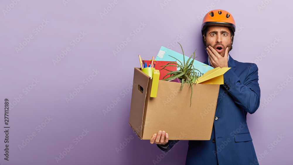 Studio shot of terrified employee holds chin and looks in terror, poses with carton container filled with stationery and houseplant, being dismissed from office, finds new job, wears expensive outfit Studio shot of terrified employee holds chin and looks in terror, poses with carton container filled with stationery and houseplant, being dismissed from office, finds new job, wears expensive outfit