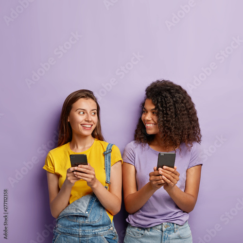 Two cute millennial girls use cell phones chatting online, find love in chat, have fun together, post new photo in social networks, addicted to modern technologies, wear casual clothes, pose indoor photo