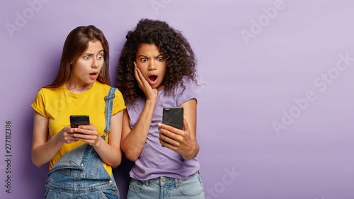 Fotografie Waist up portrait of multiethnic women stare with confusion at modern cellular, see distrubing message on screen, receive feedback from superstar, being speechless, pose indoor, blank space