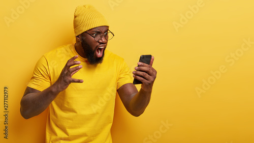 Canvas Print Outraged annoyed dark skinned man stares angrily at smart phone screen, shouts with irritation, dissatisfied with received message, has thick beard, wears casual yellow t shirt and headgear