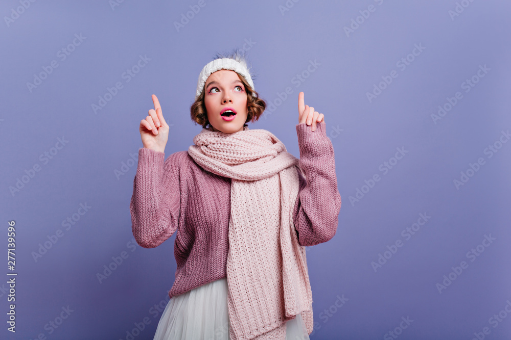 Thoughtfully short-haired girl in cute hat looking up with mouth open. Carefree female model posing in winter accessories on purple background.