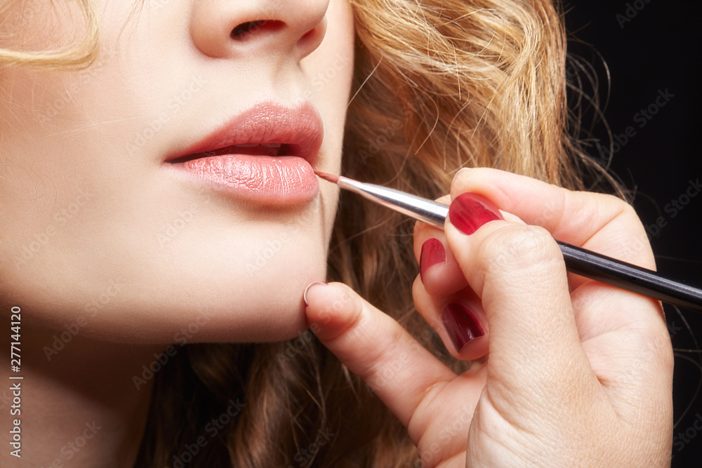 Makeup artist visagiste using brush for application lipstick on lips of young caucasian model with wavy ginger hair Makeup artist visagiste using brush for application lipstick on lips of young caucasian model with wavy ginger hair