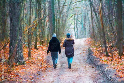 Two girls walk along a path in a park in the forest in autumn in warm clothes.