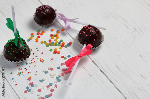 Fototapeta Naklejka Na Ścianę i Meble -  Cake pops decorated with a bow of braid. They lie on the boards, painted white. Nearby is scattered multicolored decorative topping.