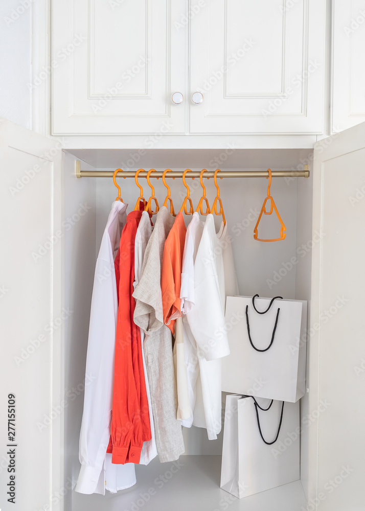 Collection of clothes hanging on rack in white wardrobe in dressing room and two paper shopping bags. Fashion shopping concept background.
