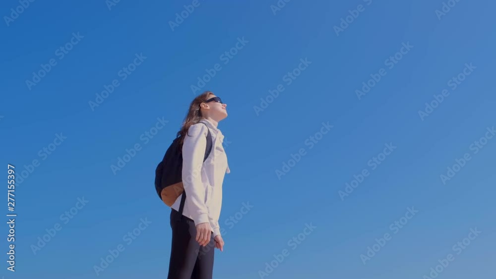Young woman traveller with backpack in sunglasses is enjoying nature on blue sky background. She is standing and looking at sky. Escape of reality. Freedom and carefree life. Young woman traveller with backpack in sunglasses is enjoying nature on blue sky background. She is standing and looking at sky. Escape of reality. Freedom and carefree life.