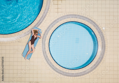 Beautiful and sexy woman have sunbathe in brown swimmsuit next to round pools, laying on a blue sunbed. Aerial model image, from birds eye view, by drone.