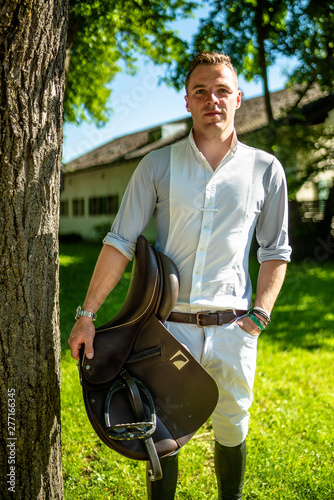 Junger attraktiver Mann in Hemd und Reithose, mit Pferdesattel in der Hand.