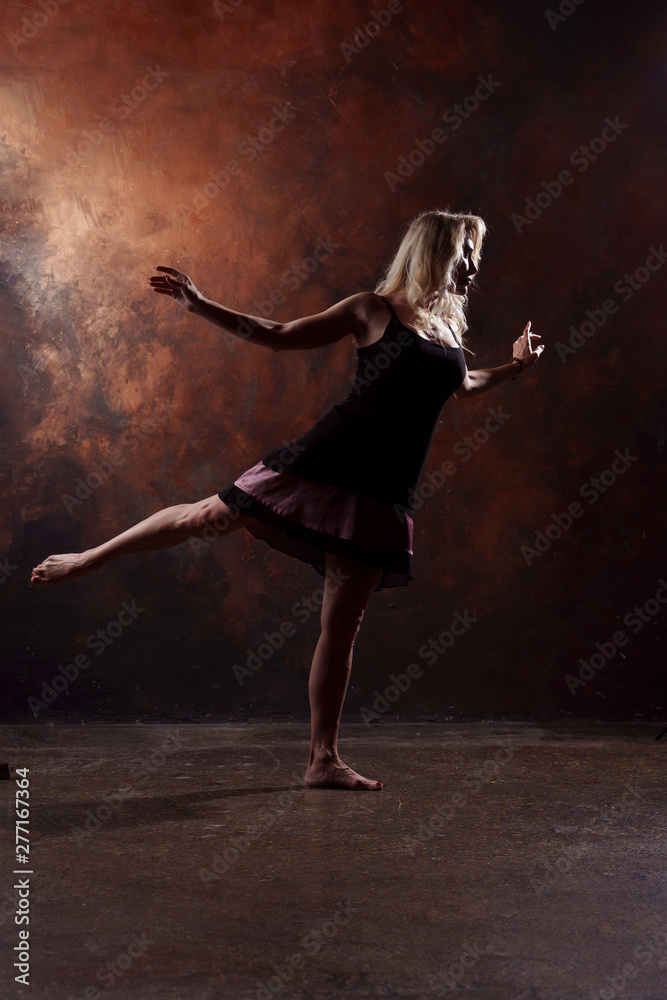 Full-length photo of dancing blonde woman looking in side in short black dress on brown background