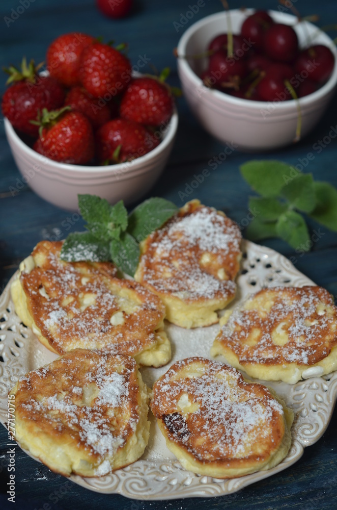 Fried pancakes on a rustic saucer sprinkled with powdered sugar. Strawberries and cherries in small bowls on a blue wooden background