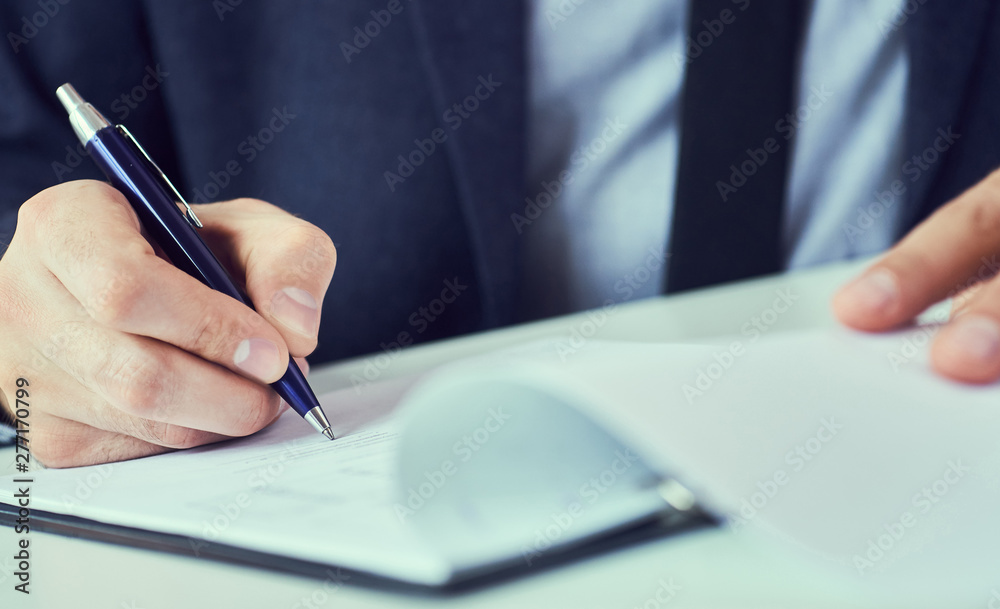 Hand of businessman in suit filling and signing with blue pen partnership agreement form clipped to pad closeup. Management training course, some important document, team leader ambition concept. Hand of businessman in suit filling and signing with blue pen partnership agreement form clipped to pad closeup. Management training course, some important document, team leader ambition concept.