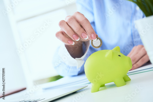 Woman's hand putting money coin in green piggy bank close-up in office background. Growing business, pension and insurance savings concept.