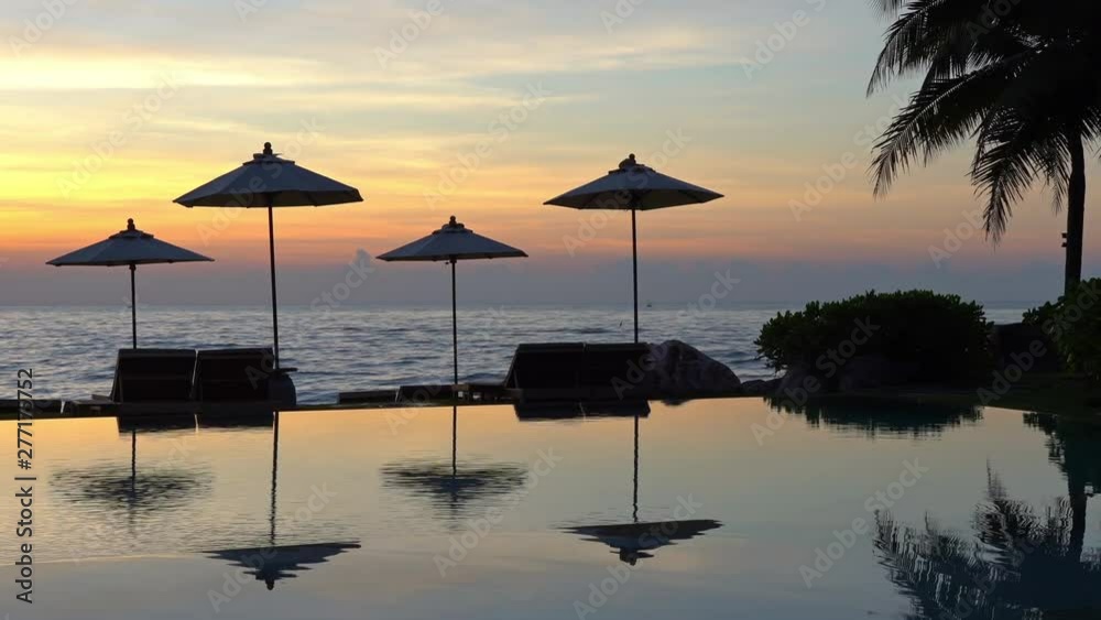 Static shot of beach sunbed loungers and umbrellas on the beach behind an infinity pool during sunset