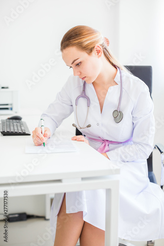 Young smiling female blonde doctor sitting at the desk on a black chair with a pen and stethoscope on her neck and wrightm something