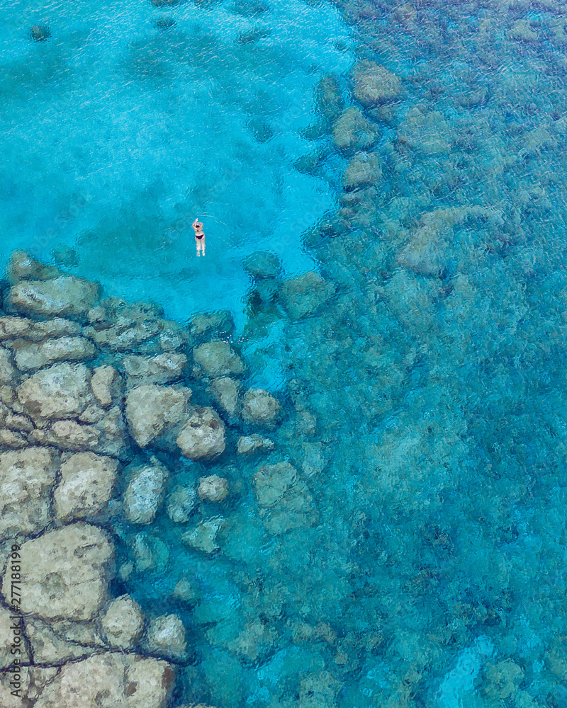 Obraz premium An aerial view of the beautiful Mediterranean Sea, with a lonely swimmer, where you can see the rocky textured underwater corals and the clean turquoise water of Protaras, Cyprus