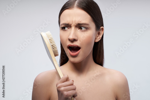 Beautiful young woman with massage brush standing against light blue background