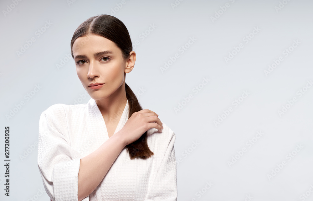 Beautiful young woman in bathrobe standing against light blue background Beautiful young woman in bathrobe standing against light blue background